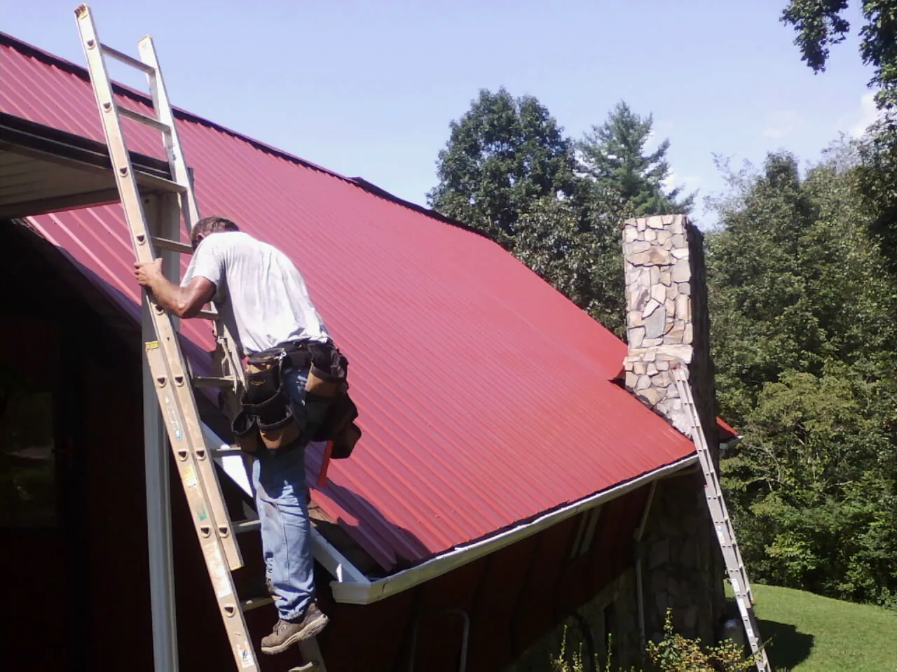 Roofer on a ladder beside a red metal roof and stone chimney — Soffit & Fascia Repair by Roofing Brothers in Porum